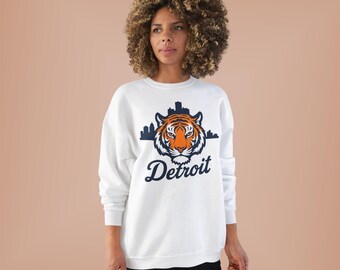 May include: White sweatshirt with a tiger graphic and the word "Detroit". The tiger is orange and black, with a blue skyline above. The sweatshirt has long sleeves. The design is in a script font.