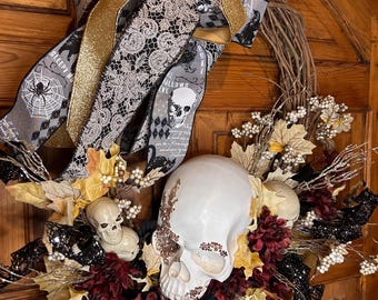 May include: A Halloween wreath with a large white skull, smaller skulls, and decorative elements. The wreath features black, gold, and gray ribbons with spiderwebs and skull motifs. Burgundy flowers and gold leaves add color.