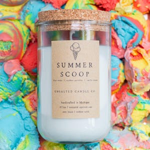 May include: A clear glass candle jar with a cork lid and a light blue wax interior. The label features an ice cream cone icon, the text "SUMMER SCOOP", "blue moon | rainbow sprinkles | vanilla cream", "UNSALTED CANDLE CO", and "10.5oz".