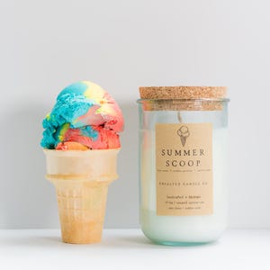 May include: A multi-colored scoop of ice cream in a tan waffle cone next to a clear glass candle jar with a cork lid. The candle label reads SUMMER SCOOP, UNSALTED CANDLE CO, and 10.5oz | coconut apricot wax | non toxic | cotton wick.