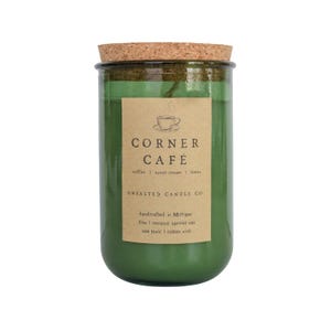 May include: A green glass jar candle with a cork lid. The label reads "CORNER CAFÉ" with the scent notes "coffee, sweet cream, Ionia." The candle is handcrafted in Michigan and made with coconut apricot wax.