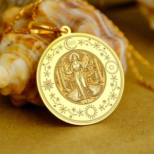 May include: A gold-colored pendant necklace featuring a detailed circular charm. The charm depicts a figure within a detailed border of stars, moons, and suns. The necklace is displayed on a seashell and a neutral background.