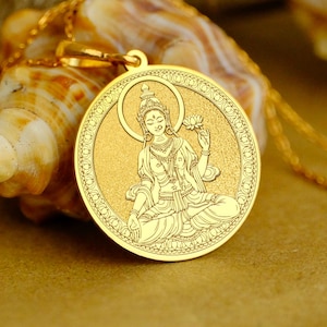 May include: A gold-colored pendant necklace featuring a detailed depiction of a seated figure within a circular frame. The pendant is suspended from a delicate gold chain. The background includes a seashell and white flowers, creating a soft, natural setting.