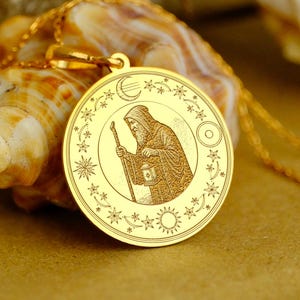 May include: Gold-colored pendant necklace featuring a detailed engraving of a robed figure with a staff, surrounded by celestial symbols. The pendant is round and hangs on a delicate gold chain. The background includes a seashell and flowers.