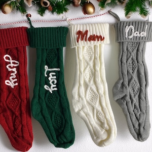 Puede incluir: Cuatro calcetines navideños de punto trenzado colgados en fila. El calcetín rojo lleva el nombre Amy, el verde el nombre Lucy, el blanco el nombre Mom y el gris el nombre Dad, bordados en el dobladillo de cada calcetín.