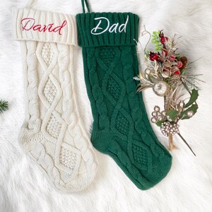 Op de afbeelding: Twee kabelgebreide kerstsokken, één crèmekleurige met "David" geborduurd in rood, en een groene met "Dad" in wit. De sokken worden gepresenteerd op een witte, pluizige ondergrond, met feestelijke bloemaccenten.