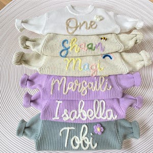 May include: A stack of colourful knitted sweaters, each embroidered with a name. The sweaters are in white, beige, lilac, and teal. Each sweater has a unique name embroidered in a contrasting colour. The sweaters are arranged on a textured, white surface.