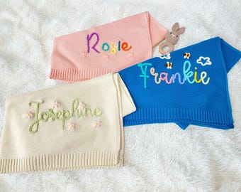 May include: Three personalised baby blankets in pink, blue, and cream, each embroidered with a name in colourful thread. The pink blanket says "Rosie", the blue blanket says "Frankie", and the cream blanket says "Josephine". A wooden bunny teether is also visible.
