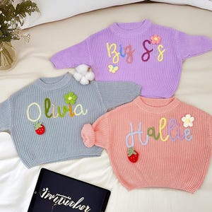 May include: Three pastel-coloured knit sweaters with embroidered names and decorative elements. One is lavender with "Big Sis", another is light blue with "Olivia", and the third is peach with "Hallie". Each has a flower and strawberry accent.