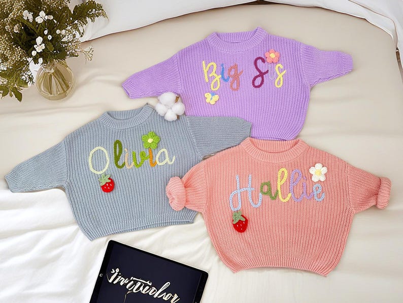 May include: Three pastel-coloured knit sweaters with embroidered names and decorative elements. One is lavender with "Big Sis", another is light blue with "Olivia", and the third is peach with "Hallie". Each has a flower and strawberry accent.