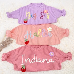 May include: Three pastel-coloured children's sweaters with embroidered names and decorative elements. The sweaters are lavender, peach, and pink, each featuring a name in cursive script, a strawberry, and a flower.