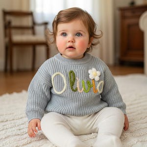 May include: A baby in a pale blue knitted jumper with the name "Olivia" embroidered in colourful thread. A white flower is also embroidered on the jumper. The baby wears white trousers and sits on a white rug.