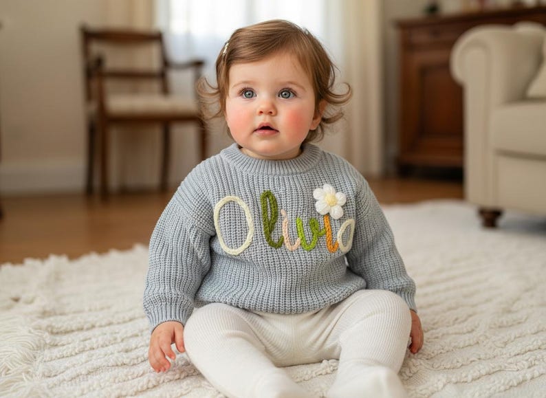 May include: A baby in a pale blue knitted jumper with the name "Olivia" embroidered in colourful thread. A white flower is also embroidered on the jumper. The baby wears white trousers and sits on a white rug.