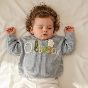May include: A pale blue baby sweater with the name "Olivia" embroidered in colourful thread, and a white flower appliqu&eacute;. The sweater is ribbed and has long sleeves. The baby is lying on a white sheet.