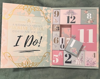 May include: A wedding countdown calendar with a pastel color scheme. The open box reveals numbered compartments, with the text "A Wedding Countdown From Me To You 12 Days Until You Say I Do!" on the left side. The numbers are in a mix of pink and white.