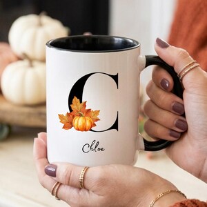 Puede incluir: Una taza de cerámica blanca con interior y asa negros, que presenta una letra C grande en negro decorada con hojas de otoño naranjas y amarillas y una pequeña calabaza. El nombre Chloe está impreso en una fuente cursiva negra bajo la letra.