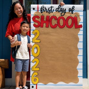 May include: A large vertical sign designed to look like a piece of notebook paper with blue horizontal lines and a red margin. The text reads first day of SHCOOL in black and red, with the year 2026 in yellow stacked vertically on the left side.
