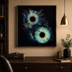 Op de afbeelding: Een vierkant ingelijst kunstwerk aan de muur met twee gedetailleerde menselijke irissen tegen een effen zwarte achtergrond. De bovenste iris toont diepblauwe en gouden tinten, terwijl de onderste iris lichtblauwe en groene tinten met stralende patronen heeft.