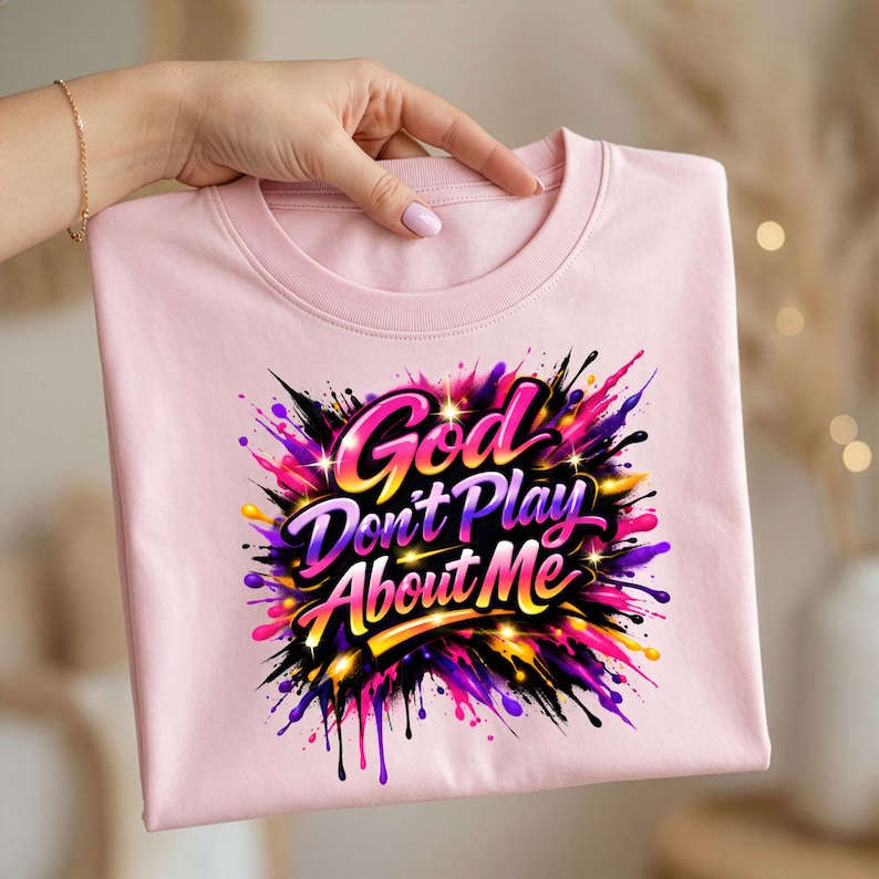 May include: A light pink crew neck t-shirt with the text "God Don't Play About Me" in a colorful, splash-style design. The graphic features shades of pink, purple, yellow, and black.