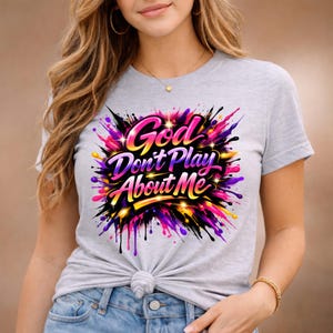 May include: A light gray t-shirt featuring the text "God Don't Play About Me" in a colorful, stylized design. The graphic includes pink, purple, yellow, and black elements, creating a vibrant, eye-catching look. The shirt is a casual wear item.