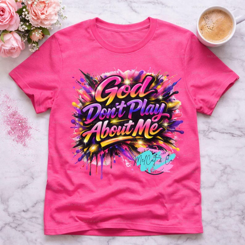 May include: A bright pink t-shirt with the text "God Don't Play About Me" in a colorful, graffiti-style design. The graphic includes splashes of purple, yellow, and black. The shirt also features the logo "McCat's & Co."