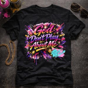 May include: Black t-shirt with the text "God Don't Play About Me" in a colorful, graffiti-style design. The design features pink, purple, yellow, and blue paint splatters. A gold chain and sunglasses are visible.