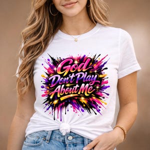 May include: White t-shirt featuring the text "God Don't Play About Me" in a vibrant, colorful design. The graphic includes shades of pink, purple, yellow, and black, creating a bold and eye-catching statement. The shirt is a casual, everyday wear item.