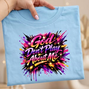 May include: Light blue t-shirt with the text "God Don't Play About Me" in a colorful, stylized design. The graphic features pink, purple, yellow, and black accents, creating a vibrant and eye-catching look. The shirt is held by a hand.
