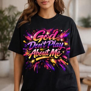May include: Black t-shirt with the text "God Don't Play About Me" in a colorful, stylized design. The shirt features a vibrant explosion of pink, purple, yellow, and orange colors. The words "Bold Faith Streetwear" are at the top.