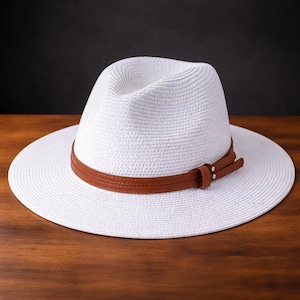 May include: A white fedora hat with a brown leather band. The hat has a wide brim and a classic shape. The hat is sitting on a wooden surface.
