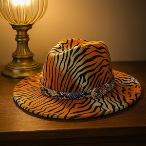 Può includere: Un cappello fedora con un vivace motivo a strisce zebrate arancioni e nere. Il cappello presenta una fascia decorativa con una stampa a pelle di serpente nei toni del blu, marrone e bianco. Il cappello è appoggiato su una superficie di legno.