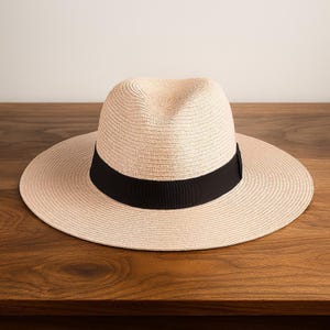 May include: A light beige straw fedora hat featuring a wide brim and a solid black grosgrain ribbon band encircling the crown. The hat has a pinched centre crease and a structured, woven texture throughout the entire surface of the material.