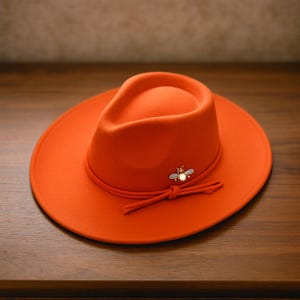 Op de afbeelding: Fel oranje fedora hoed met een bijvormige broche. De hoed heeft een brede rand en een oranje band die in een strik is geknoopt. De hoed is gemaakt van een zacht materiaal.