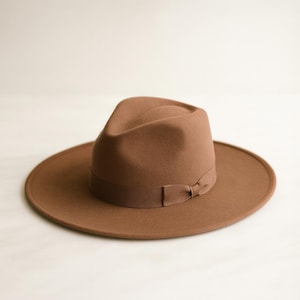 May include: A brown fedora hat with a wide brim and a matching ribbon band tied in a bow. The hat has a classic, structured shape and is made of a soft, felt-like material. The hat's brim measures approximately 3 inches.