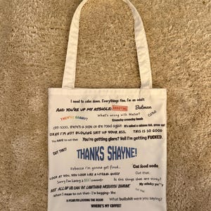 May include: A cream-coloured canvas tote bag featuring a variety of printed phrases and quotes in different fonts and colours, including the prominent blue text THANKS SHAYNE! and other phrases like I need to calm down, Batman, and WHERE'S MY COFFEE?