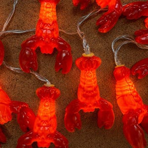 May include: A string of red lobster-shaped lights with a warm orange glow. The lights are strung on a thin, silver wire, and are arranged on a textured, brown surface. The lobster lights are a fun and festive decoration.