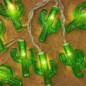 May include: A string of green cactus-shaped fairy lights with a warm glow. The lights are strung on a clear wire and are arranged on a textured, tan surface. The cactus lights are a fun and festive decoration.