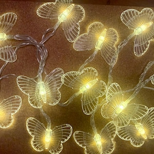 Pode incluir: Uma sequência de luzes transparentes em forma de borboleta com um brilho amarelo quente. As borboletas são feitas de um material translúcido e são conectadas por um fio fino prateado. As luzes estão dispostas sobre uma superfície marrom.