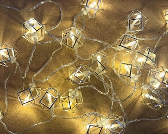 Könnte beinhalten: Eine Lichterkette mit dekorativen, würfelförmigen Lampenschirmen. Jeder Schirm hat einen silbernen Metallrahmen und enthält ein warmes, gelbes Licht. Die Lichter sind auf einem dünnen, transparenten Draht aufgereiht und erzeugen eine gemütliche und moderne Ästhetik.