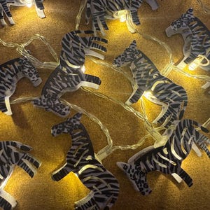 Op de afbeelding: Decoratieve lichtslinger met zebra-vormige uitsparingen met zwart-witte strepen. De zebra's worden verlicht door kleine, warmwitte lampjes. De lichten zijn gerangschikt op een getextureerd, goudbruin oppervlak.