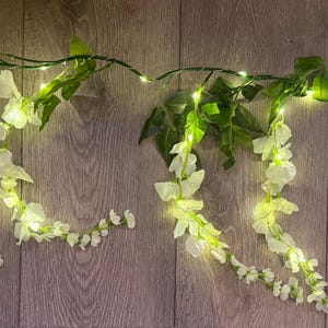 Puede incluir: Guirnalda decorativa con flores blancas y hojas verdes, iluminada por pequeñas luces LED blancas cálidas. La guirnalda está dispuesta sobre una superficie de madera, creando un ambiente suave. Ideal para decoración del hogar o eventos.