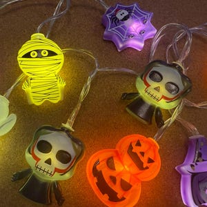 May include: A string of Halloween lights with various shapes including ghosts, mummies, pumpkins, and skulls. The lights are in vibrant colours, such as orange, yellow, and purple, creating a festive and spooky atmosphere. Ideal for Halloween decorations.