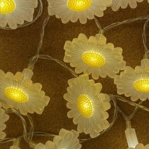 May include: String lights with daisy-shaped bulbs, emitting a warm yellow light. The bulbs have translucent white petals and bright yellow centres, creating a cheerful atmosphere. Ideal for decorating homes or for special occasions.