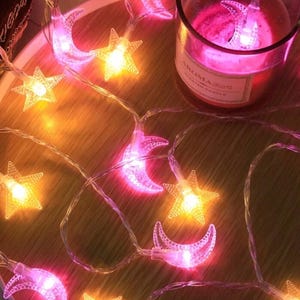 May include: A string of decorative lights featuring illuminated crescent moons and stars. The lights are strung on a clear wire and emit a warm, inviting glow. A candle in a glass jar is also present, adding to the ambiance.