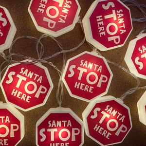 May include: A string of Christmas lights featuring red and white stop sign-shaped lights. Each sign reads "SANTA STOP HERE" in white lettering. The lights are connected by a clear wire, creating a festive holiday decoration.