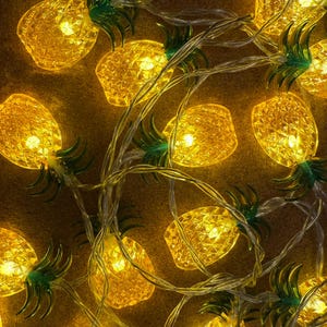 May include: String of decorative lights featuring pineapple-shaped bulbs. The lights are yellow and emit a warm glow. Each pineapple has green leaves at the top. The string is clear, and the lights are arranged closely together.