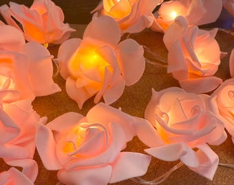 May include: A string of soft pink rose-shaped lights. Each rose has a warm yellow glow emanating from its center. The lights are strung together on a thin, clear wire, creating a delicate and romantic ambiance. Ideal for home decor or special events.