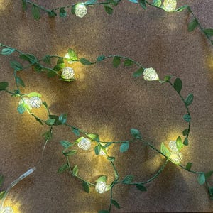 May include: A string of decorative lights featuring small, white, glowing flower-shaped bulbs. The lights are strung along a green vine with small green leaves. The lights are on a brown surface.