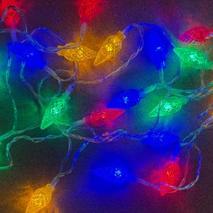 May include: A string of colorful Christmas lights with red, green, blue, and yellow bulbs. The lights are arranged in a tangled pattern, with the bulbs illuminated against a dark background. The bulbs have a textured, faceted design.