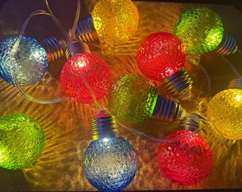 Puede incluir: Una cadena de luces LED decorativas con globos de vidrio texturizado multicolor en rojo, azul, verde y amarillo. Cada bombilla esférica tiene una base de rosca metálica y está conectada por un cable eléctrico transparente.